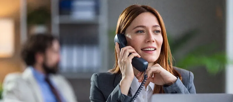 A woman in a grey blazer smiles as she answers a call on a desk phone, with a blurred colleague in the background